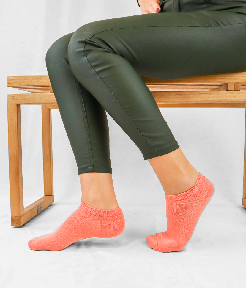 Egy lány lábait egymás mellett tartva egyszínű, barackvirág Goisocks bokazoknit visel, miközben ül egy padon.